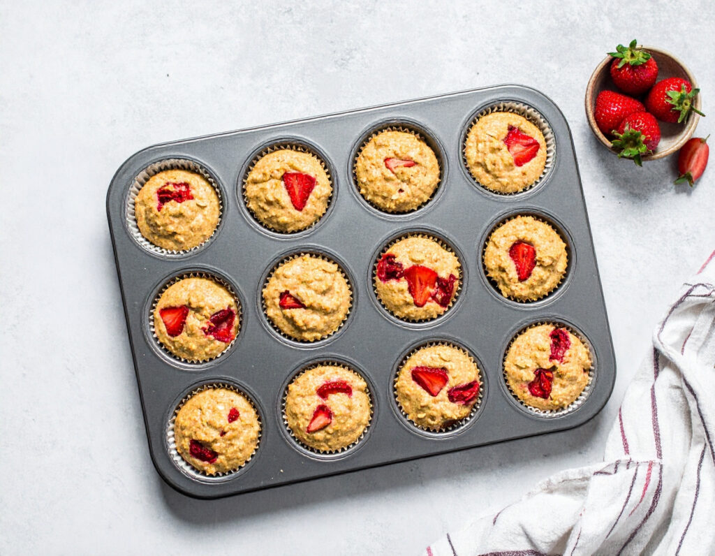 Ungebackene Erdbeermuffins im Muffinblech mit sichtbaren Erdbeerstücken, daneben Schale mit frischen Erdbeeren