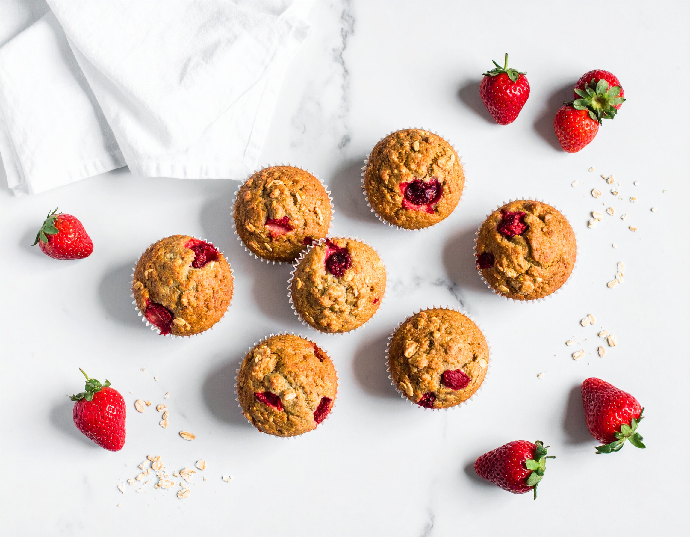 Acht goldbraune Erdbeermuffins von oben auf Marmor, garniert mit frischen Erdbeeren und Haferflocken